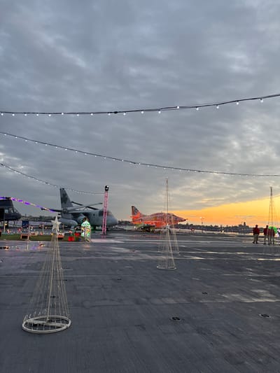 The image presents a festive scene on the flight deck of an aircraft carrier, likely the USS Midway Museum in San Diego, during the transition from dusk to evening. The sky overhead is heavily overcast with grey clouds, but a vibrant orange and yellow sunset is visible on the horizon, casting a warm glow over a distant city skyline and a body of water.
The main subjects are the Christmas decorations and the aircraft on the deck. Strings of white lights are strung across the top of the frame, creating an overhead canopy. Several large, conical light sculptures, made of white frames and embedded lights, are scattered across the deck, one in the foreground topped with a white star.
Two aircraft are prominent: a fighter jet, fully covered in bright orange and red Christmas lights, is positioned towards the rear of the deck. Closer to the foreground and to the left, a large helicopter, possibly a CH-46 Sea Knight, is also decorated, with a festive, illuminated gift box-like structure placed near its base.
In the far background, two silhouetted individuals are visible on the deck, observing the sunset. The scene depicts a holiday event, transforming the military vessel into a public attraction with seasonal illuminations. The time of day is clearly sunset or early twilight, creating a dramatic and atmospheric setting. The weather appears cloudy, with a distinct break in the clouds at the horizon for the sunset. No visible text is present in the image.