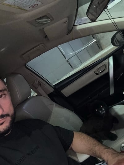 The image shows the interior of a car, likely a sedan, with a person and a black cat. The person is in the driver's seat, partially visible, wearing a black t-shirt. A black cat is in the passenger seat, looking towards the camera with its eyes reflecting light. The car appears to be parked, with the sunroof open, revealing a glimpse of the sky and an exterior environment that does not provide definitive clues about the specific location within Mai Khao, Thailand. The interior is dimly lit, typical of a car's interior at night or in a shaded area. The upholstery is a light neutral color, and car interior elements like the rearview mirror, door handle, and seatbelt are visible. There are no other people or significant objects that would indicate a specific urban setting within Mai Khao.