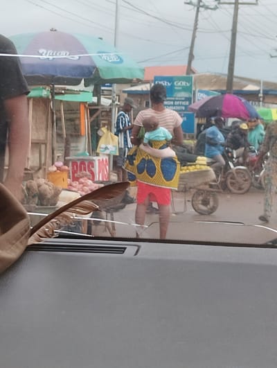 The image depicts a bustling street scene in Aketu-Oja, Nigeria, viewed from a vehicle. A woman, her back to the camera, carries a baby wrapped in a colorful cloth around her torso. She wears red shorts and a striped t-shirt. In the background, a stall displays produce, including onions and what appear to be yams. A sign with "POS POS" written in red is visible. To the left, a large umbrella displays "Hugo" and "Wazo". Several other individuals and motorcycles are present, suggesting a lively market or transport hub. The overall environment appears to be an outdoor commercial area with overhead wires and electricity poles.