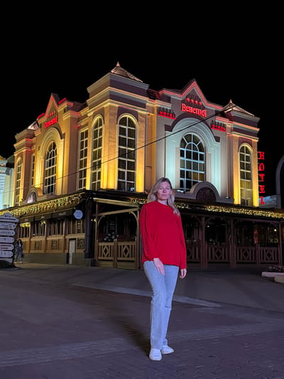 A woman stands in front of a brightly lit hotel at night in Kislovodsk, Russia. The hotel, named "Венеция" (Venetsiya), is a prominent building with illuminated red lettering and decorative lights. The woman, dressed in a red sweater and light-colored jeans, is facing the camera with her eyes closed. The street in front of the hotel is paved with cobblestones. A signpost with various inscriptions is visible to the left of the frame. The environment is dark, illuminated primarily by the hotel's lights.