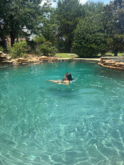 A woman is swimming in an outdoor swimming pool. The pool is surrounded by trees and landscaping. The scene is located in Southlake, United States of America.