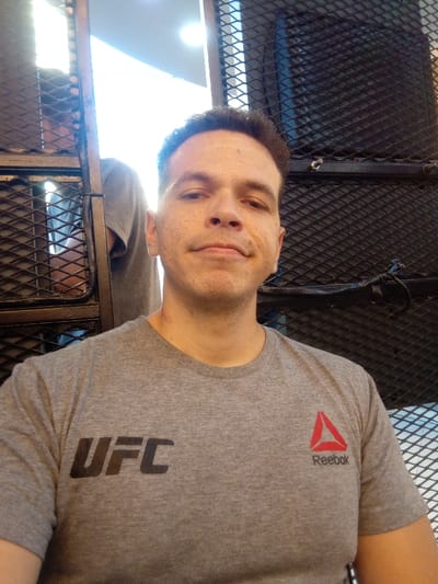 The image displays an indoor scene, situated in Quilmes, Argentina, featuring a male subject in the foreground. The subject has short dark hair, light-to-medium skin tone, and visible freckles or stubble on his cheeks and jawline. He is looking directly at the camera with a neutral expression. He wears a grey short-sleeved t-shirt with "UFC" printed in black on the left chest and a red Reebok logo with "Reebok" text on the right chest. A small, reddish mark is visible on the right side of his neck near the collarbone.
The background consists of dark, diamond-patterned metal mesh structures. On the left side, behind a dark vertical beam of this structure, a bright, overexposed light source is present. Partially visible through the mesh and behind this light is the torso of a second individual, wearing a grey t-shirt, consistent in color with the main subject's attire. The right side of the background shows a darker expanse of the same diamond-patterned metal mesh, with some dark cables or objects integrated into the structure. The environment suggests a facility with industrial design elements, possibly a gym or a public venue.