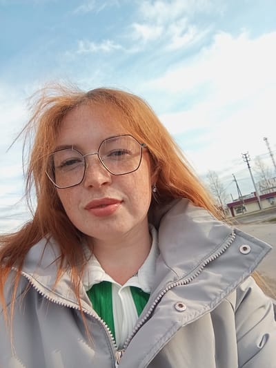 The image shows a woman with red hair and freckles wearing glasses, a white top with a green collar, and a gray jacket. She is outdoors, and the background reveals a blue sky with light clouds and a street scene. A building with a red roof and white walls is visible in the background. There are utility poles and bare trees lining the street. Based on the image context, the location is likely Yurgamysh, Russia.
