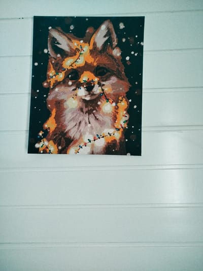 A painting of a fox wearing Christmas lights is hanging on a wall made of horizontally paneled wood. The painting is dark with a black background, and the fox is depicted in warm tones of orange, brown, and white. Snowflakes are scattered around the fox. The Christmas lights are a mix of yellow, red, green, and blue. The wall is painted white and has a slightly textured appearance due to the wood paneling. There are no people or other objects visible in the frame. The image appears to be taken indoors.