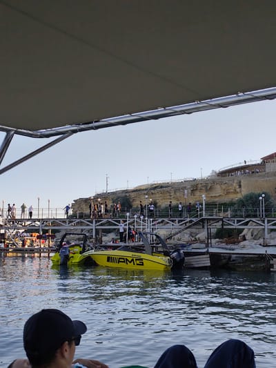 The image depicts a lively scene at a marina, likely in Aktau, Kazakhstan. The foreground shows the legs and back of a person wearing a black baseball cap, seated and looking out towards the water. The focus of the image is the marina, where several boats are docked.
A prominent yellow speedboat with the "AMG" logo and "MAN 0330" text is the most striking object. Another, similar yellow boat is docked next to it. The boats are in the water, with a wooden dock visible behind them.
On the dock and a walkway extending over the water, a group of people are gathered. They appear to be casually strolling and observing the surroundings. The walkway is elevated, providing a view of the marina and the cliffs in the background.
Behind the walkway, a rocky cliff face rises, with a building or structure situated on its summit. The cliff has a weathered, sandy appearance. Scattered vegetation grows on the cliff and around the marina area. The sky is clear and a pale blue, indicating it is likely daytime, possibly late afternoon or early evening given the soft light. The overall atmosphere seems relaxed and leisurely, with people enjoying a day at the waterfront.
