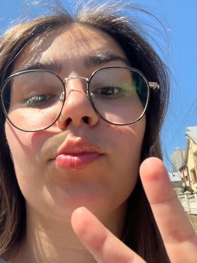 This is a close-up, upside-down selfie of a young woman wearing glasses. She has dark hair and is making a peace sign with her right hand, which is partially obscuring her face. Her lips are slightly parted, and she is wearing glossy pink lipstick. The image appears to be taken outdoors on a sunny day, as indicated by the bright blue sky and the strong sunlight reflecting off her skin and hair. In the background, there are buildings that suggest an urban or town setting. The reflection in her glasses shows a building with a light-colored facade and windows, as well as a blue sky. The overall mood of the image is casual and playful, fitting for a selfie. The location context provided indicates Chahor, Ukraine.