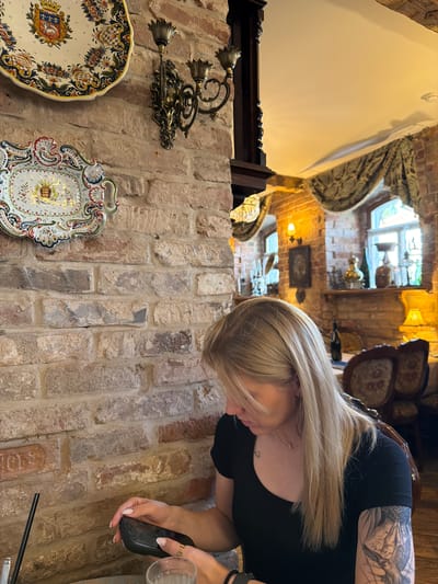 A blonde woman with a tattoo on her arm sits at a table in a restaurant in Suzdal, Russia. She is looking down at her phone, which she holds in her hands. The restaurant has exposed brick walls, ornate decorative plates on the wall, and antique-style furniture. Sunlight streams in through a window in the background.
