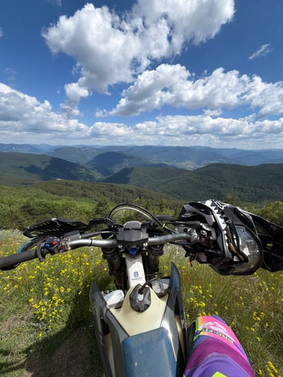 A first-person perspective shot captures a dirt bike parked on a grassy, wildflower-covered slope with a vast, mountainous landscape in the background under a blue sky with scattered white clouds. The handlebars of the bike are visible in the foreground, equipped with a helmet resting on the right side. A person's legs, wearing brightly colored motocross pants, are also visible in the lower right corner. The scene appears to be a scenic overlook in a rural or mountainous region.