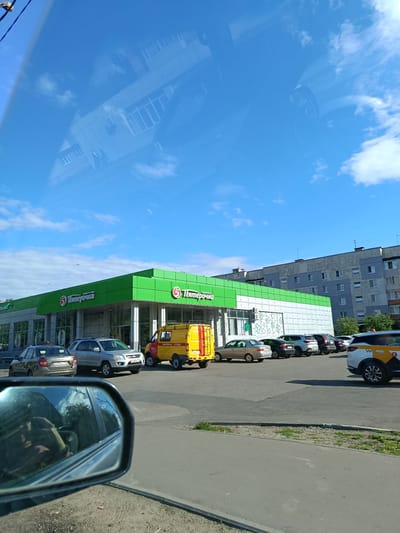 This media file captures an outdoor scene in Orekhovo-Zuyevo, Russia, on a bright, sunny day. The foreground shows a car's side mirror reflecting the interior of the vehicle and the sky with some clouds. In the mid-ground, a bright green and white "Pyaterochka" supermarket is visible. Several cars are parked in front of the store, including a silver SUV, a dark sedan, and a yellow van with "AVARIKAYNAYA SLUZHBA" (Emergency Service) written on its side. A white taxi with yellow and black markings, featuring the "Я" symbol, is also present. The background consists of apartment buildings. The scene appears to be during the daytime, given the bright sunlight and clear blue sky. The overall atmosphere is calm and ordinary, suggesting a typical day in a residential area with a local shopping center. Visible text includes "Pyaterochka" on the supermarket facade, "AVARIKAYNAYA SLUZHBA" on the yellow van, and license plate information on some of the cars.