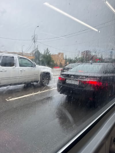 The image depicts a traffic scene in Zelenograd, Russia, on a rainy day. The view is from inside a vehicle, looking out through a wet window. In the foreground, a black Toyota Camry is visible with its brake lights illuminated. The license plate reads "H223YT 777 RUS". To the left of the Camry, a white SUV, possibly a Chevrolet Suburban, is seen in traffic. The road surface is wet and reflects the car lights and surrounding environment. In the background, buildings, trees, and utility poles are discernible through the rain and window. The sky appears overcast.