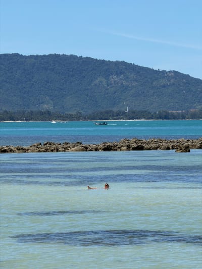 The image depicts a serene coastal scene in Baan Thong Krut, Thailand. The composition is dominated by a lush, verdant mountain slope that recedes into the distance on the left side of the frame. The mountain is densely covered with trees, suggesting a tropical environment.
The ocean occupies the central and right portions of the image. The water is a vibrant turquoise, with clear visibility revealing streaks of lighter and darker blue, likely due to variations in depth and the play of sunlight on the seabed. A rocky outcrop juts out from the sea, creating a textured contrast with the smooth water.
In the foreground, on the right side of the image, a person is partially submerged in the water, appearing to be swimming or wading. They are looking towards the left, with only their head and upper body visible. The water is calm, with gentle ripples reflecting the sunlight.
Further out in the ocean, several boats are visible. One is a larger vessel with a dark hull, while others appear to be smaller, possibly traditional Thai longtail boats. The presence of these boats suggests a location frequented by tourists or locals engaged in water activities.
The sky is a clear, bright blue, indicating a sunny day with no clouds. The time of day appears to be midday or early afternoon, given the intensity of the sunlight. The overall atmosphere is peaceful and idyllic, capturing the essence of a tropical beach holiday. There is no visible text in the image.