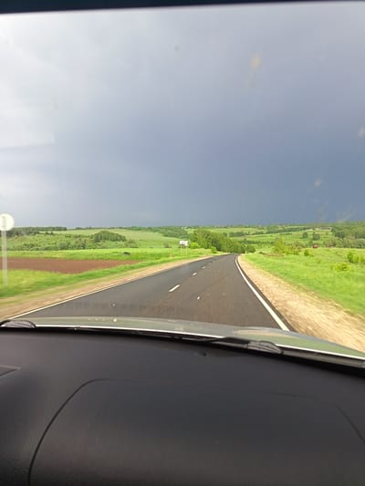 Captured from inside a vehicle moving along a paved road, the image shows a view towards the horizon in what appears to be a rural setting. The road has white line markings. Adjacent to the road are green fields, interspersed with trees and vegetation. The sky is overcast with dark storm clouds dominating the upper portion of the frame. The dashboard and a windshield wiper of the vehicle are visible in the foreground. Based on the location Yemeshevo, Russia, the environment is likely in that geographic region.