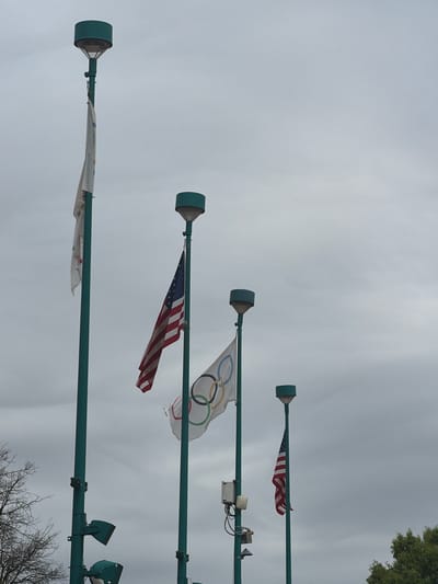The image shows a series of flagpoles against a cloudy sky. From top to bottom, the flags appear to be: an American flag, the Olympic flag, another American flag, and a fourth faded and unidentifiable flag. The presence of the Olympic flag and the flags is indicative of the Atlanta area due to the Centennial Olympic Games. The flags are likely displayed in Atlanta, Georgia, United States.