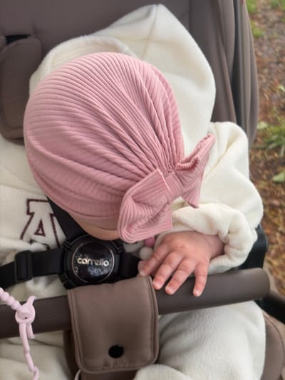 A top-down, medium shot depicts an infant seated in a stroller. The infant is wearing a white, textured jacket and a pink, ribbed knit hat that resembles a tied bundle. The baby's hand rests on the stroller's padded bar, near a black wristwatch with the word "Carrello" visible on the watch face. A brown pouch is affixed to the stroller's bar. A portion of the stroller's brown seat and frame are visible. Pink stroller clips are knotted around the bar. The location cannot be determined from the image.