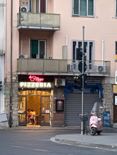 The image depicts a street view in Pisa, Italy, focusing on a pizzeria named "Da Filippo Pizzeria." The pizzeria has a prominent neon sign and a large glass storefront, through which two individuals are visible sitting at a table. The building facade is light pink, with balconies on the upper floors. Adjacent to the pizzeria is a store offering "Luggage Storages," "SIM CAR," and "Money Transfer" services, indicated by signs. A traffic light stands near the entrance of the luggage storage establishment. A pink scooter and a bicycle are parked on the sidewalk in front of the businesses. The street in the foreground is paved and appears to be an intersection.