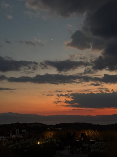 The image captures a serene twilight scene in the Municipal Unit of Kranidi, Greece, likely from a vantage point overlooking the Argolic Gulf. The sky is a canvas of gradient hues, transitioning from a deep indigo at the top to a vibrant orange and red near the horizon, punctuated by dramatic, dark clouds. A faint crescent moon is visible high in the sky.

In the foreground, a dimly lit outdoor seating area is set amidst lush, dark vegetation and white flowers, some of which are illuminated by soft, warm lights. The setting suggests a restaurant or a private terrace. Two silhouetted figures are seated at a table, one with a glass of white wine, seemingly engaged in conversation or enjoying the view. Another figure stands in the mid-ground, facing away from the camera.

Beyond the immediate foreground, rolling hills covered in dark foliage form the landscape. In the distance, a range of mountains is silhouetted against the colorful sky, with the calm waters of the sea visible between them. The overall atmosphere is one of tranquility and natural beauty, typical of the coastal regions of the Peloponnese.