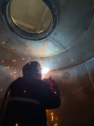 A person wearing welding safety gear is welding inside a large metal structure. The structure appears to be a section of a large pipe or tank, possibly under construction or repair. There is an opening on the left side with a circular flange attached. Welding sparks are visible at the point of contact. The location and city of Tomba, Montenegro are not discernible from the image.