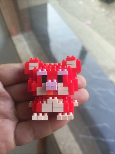 A human hand, primarily the palm and thumb, is depicted holding a small, red block-built figure resembling a cat or fox. The figure is constructed from interlocking mini-blocks in red, with white accents for the snout, inner ears, and paws, black for the eyes, light purple for the cheeks/nose bridge, and a small orange piece for the nose. The figure faces forward, supported from below by the hand. The background, blurred, reveals an indoor environment with a reflective surface, likely a tiled or wooden floor, displaying varying shades of gray and brown with rectangular patterns. A strong light source from the upper left casts a reflection on this surface. The scene is located in Pasir Muncang, Indonesia.