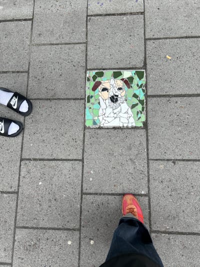 The image shows a street scene in Malmö, Sweden, at the location of 55.594 latitude and 13.011 longitude. The scene features a mosaic depiction of a white dog with brown ears set into the grey pavement. The dog is looking directly at the camera. A pair of white socks and black Nike sandals are partially visible in the bottom left of the image. A person wearing red sneakers and blue jeans is walking past the mosaic. The person is only partially visible. A small piece of trash is visible in the upper right-hand corner of the image. The pavement is made of concrete and is cracked in places. There are light-colored spots and markings scattered across the pavement. The mosaic image is in a central position on the pavement.
