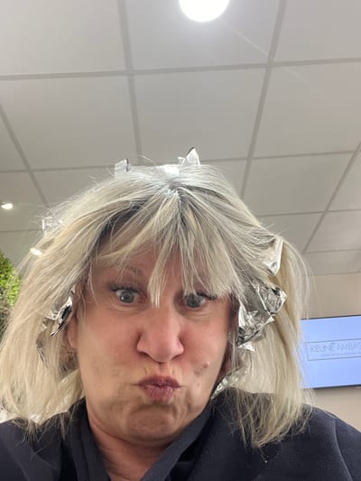 A woman with blonde hair and foil highlights is puckering her lips, seemingly taking a selfie indoors. The background features a grid-patterned white wall, suggesting a salon setting. A sign with the word "KEUNE AMBAS" is partially visible. A light fixture is present in the background. The location, Montrond-les-Bains, France, could be associated with the salon in the picture.