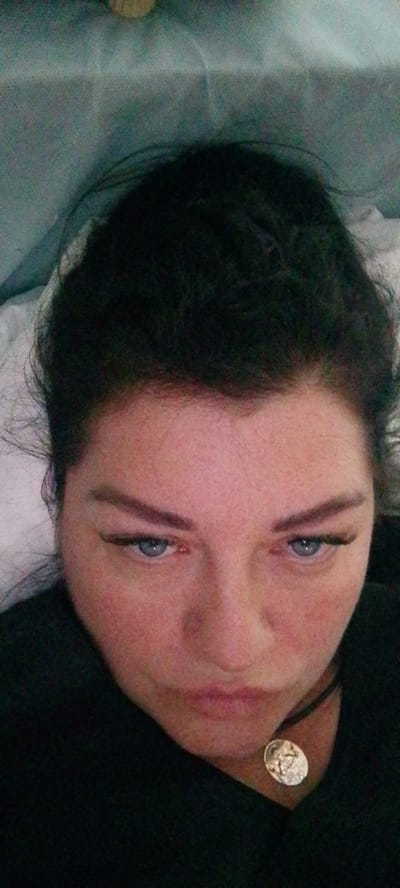 This is a close-up, overhead, selfie-style photograph of a woman with dark hair pulled up. She is wearing a black top and a pendant necklace. Her face is flushed, and she has light blue eyes with dark eyeliner and false eyelashes. Her lips are slightly parted. The background is a textured, light blue fabric, possibly bedding or upholstery. The image quality is poor, with low resolution and visible digital noise. It is not possible to determine the specific location within Volzhsky, Russia, based on the provided visual information.