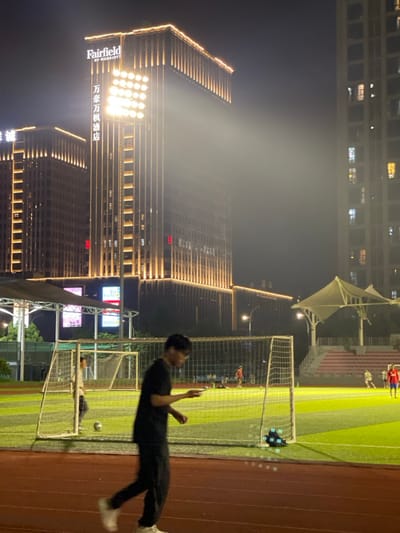 The image shows a man walking on a red running track at night. He is wearing a black shirt and black pants. He is looking down at his phone. In the background, there is a soccer field, soccer goal, and a large building with the sign “Fairfield by Marriott” illuminated with bright white lights. There are also people playing soccer on the field. The coordinates 30.177, 120.143 are in the city of Hangzhou, China. The scene is likely a sports complex within the city. The image was taken at night. The lighting is bright and the background is slightly hazy.