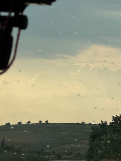 The image depicts a landscape scene viewed from inside a vehicle, as evidenced by the blurred foreground elements which appear to be parts of the car's interior, possibly a dashboard or camera equipment. The perspective is rotated, with the sky occupying the left portion of the frame and the ground and trees on the right.
The sky is a muted blue and yellow, with wispy clouds suggesting fair weather, possibly during dawn or dusk due to the soft, diffused light. Scattered across the sky are numerous small, circular, out-of-focus spots, which could be water droplets or dust on the vehicle's window or lens.
The right side of the image reveals a green, rolling landscape, likely a field or hillside. A line of trees runs along the crest of the hill, appearing as dark silhouettes against the lighter sky. In the distance, more trees and vegetation are visible, and a road or path can be discerned running through the landscape. There's a small circular sign with some red and white markings on one of the trees on the right side.
The overall impression is one of a tranquil, rural setting, possibly during a drive through the Bulgarian countryside near Tutrakan. The time of day is likely early morning or late afternoon, indicated by the soft lighting and the colors of the sky. No people or specific activities are clearly identifiable, making it a more general scenic view. The image captures a moment of quiet observation from within a moving vehicle.