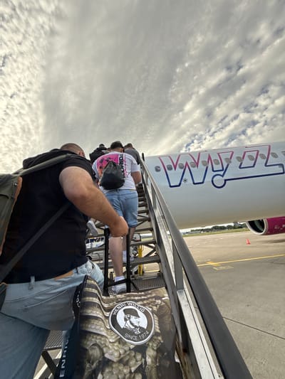 A group of people are boarding an airplane via a set of mobile stairs. The airplane is white with a purple and pink logo that reads "WIZZ". The sky is overcast with scattered clouds. The scene appears to be an airport tarmac, as indicated by the presence of the plane, stairs, and runway markings. The location is specified as Künersberg, Germany. The people are dressed casually, with some carrying bags. The perspective is from behind the group, looking up the stairs towards the airplane. One person in the foreground is wearing a black t-shirt and jeans, carrying a backpack. Another person further up the stairs is wearing a white t-shirt with a graphic and denim shorts, also carrying a backpack. The image captures a moment of transit, with the passengers in the process of boarding the aircraft.