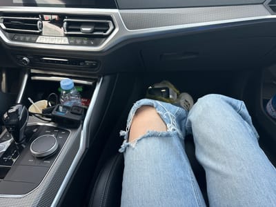The media file captures the interior of a modern car, likely a BMW, from a first-person perspective, focusing on the passenger side footwell and parts of the dashboard and center console. A person's lower body is visible, clad in light blue, distressed jeans with a large tear at the knee, and light-colored sneakers, suggesting they are seated in the passenger seat.
The scene takes place inside the car, showing details consistent with a contemporary vehicle. The dashboard features air vents, climate control buttons, and a hazard light button. A blue ambient light strip runs along the textured silver trim above the glove compartment. The center console displays the gear shifter, an infotainment control knob, and various buttons.
Notable objects include a black GoPro camera (likely a Hero model) connected by a cable, a plastic water bottle with a blue cap, and a white cup, all placed in the console area near the gear shifter. The lighting appears bright, indicating it is daytime. The location context provided is Madrets, Bulgaria.
Visible text includes numerous labels on the car's controls: "MODE", "P", "OFF", "START ENGINE", "M/S D", "REAR", "AUTO", "MENU A/C", "MAX A/C", "MEDIA", "HOME", "MAP", "NAV", "BACK", and "OPTION". The GoPro logo is also identifiable on the camera. A partially visible label on the water bottle reads "VIN".