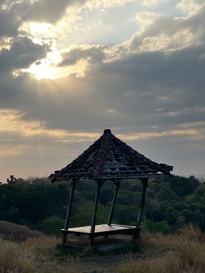 The image shows a dilapidated open-air pavilion on a grassy hilltop overlooking a dense forest. The sky above is filled with dramatic, textured clouds, with bright sunlight filtering through in rays, suggesting either sunrise or sunset. The pavilion itself is constructed of dark wood and has a tiled roof that appears to be partially collapsed or missing tiles, revealing the underlying structure. A wooden platform is visible within the pavilion. The foreground is covered in dry, tall grass, indicating a natural, perhaps somewhat overgrown, environment. The background features a thick canopy of green trees that descends down a slope, leading to what appears to be a distant body of water or coastline. The overall scene evokes a sense of isolation and natural beauty, with the decaying pavilion adding a touch of melancholy or historical resonance to the landscape. The specific location in Kubutambahan, Indonesia, implies a tropical or subtropical setting, which is consistent with the lush greenery and the appearance of the sky. There are no people or animals visible in the image.