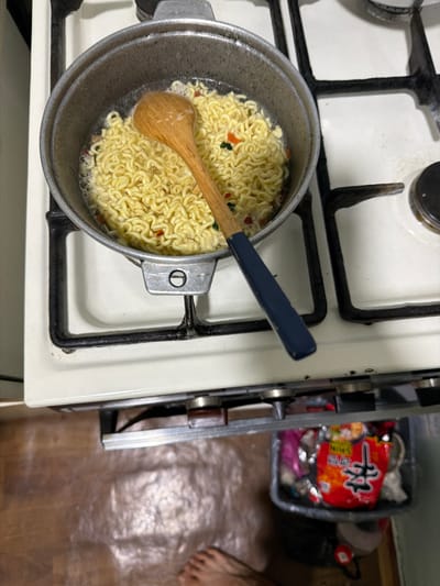 A close-up, overhead view captures instant noodles cooking in a pot on a gas stove. The pot, made of a dark, worn metal, sits on a white stovetop with black grates. Inside the pot, yellow curly noodles are submerged in boiling water, with small flecks of red and green vegetables visible. A wooden spoon with a blue handle rests within the pot, partially submerged in the noodles. The stovetop appears to be in a kitchen setting, with hints of a tiled floor and cabinet beneath. The overall scene suggests a casual, everyday meal preparation. The location is specified as Yangiiul, Uzbekistan.