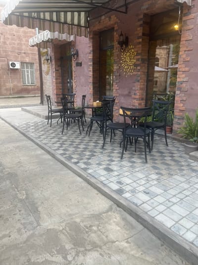 The image shows an outdoor seating area with multiple black metal chairs stacked or arranged around tables. The setting appears to be a cafe or restaurant with brick walls and a tiled floor. A section of the wall features a circular yellow sun-like decoration. The overall scene is empty, suggesting either early morning, late evening, or that the establishment is closed. The lighting is dim, possibly indicating an overcast day or evening. The ground outside the tiled area is concrete. In the background, part of a building with windows is visible, along with some greenery. The image is taken from a slightly elevated angle, looking downwards.