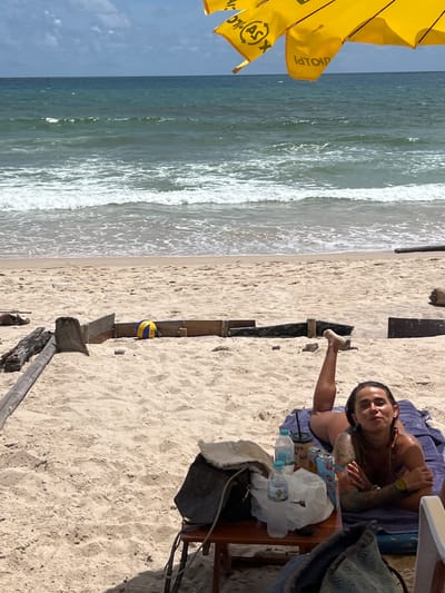 A woman is lying on a blue towel on a sandy beach, facing the camera with her legs extended. She has tattoos on her arms and is wearing a bikini top. In front of her is a small wooden table with drinks and bags. A yellow umbrella is visible at the top of the frame. The ocean and waves are in the background. The scene is set on a sunny day.