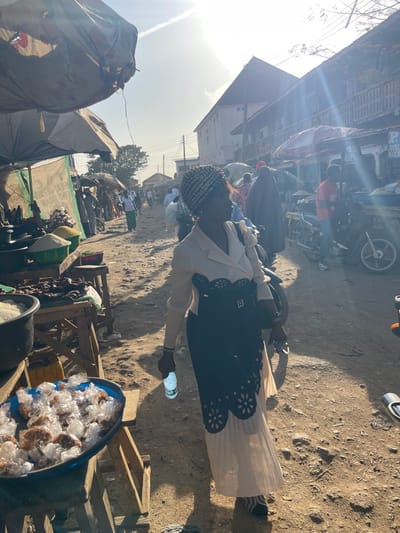 A woman wearing a light colored skirt, dark patterned top, and cream-colored jacket stands on a dirt road, holding a water bottle. She is situated in what appears to be a crowded outdoor market or street. Vendors' stalls are visible, one with items packaged in clear plastic. Motorbikes and other vehicles are parked alongside the road, with buildings and more activity in the background. The scene is bathed in bright sunlight.