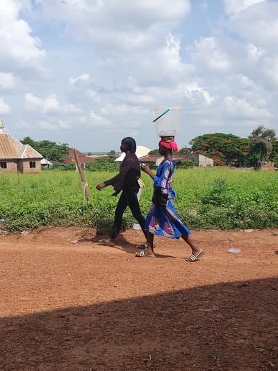 Two women walk along a reddish-brown dirt path in a rural or semi-urban setting in Idah, Nigeria. The woman in front carries a clear plastic container, possibly for water, balanced on her head. She wears a red headwrap, a vibrant blue dress with a pink and white pattern, and striped flip-flops. A small black patterned bag hangs from her waist. Behind her, the second woman walks in dark clothing, consisting of a black head covering, a dark long-sleeved top, and dark trousers, also wearing striped flip-flops.
To their left, beyond the path, is an expanse of lush green vegetation, including tall grasses and bushes. In the background, simple, single-story buildings with corrugated iron roofs are visible amongst trees. A prominent tree with reddish-orange blossoms (likely a Flamboyant tree) stands further back.
The sky is partly cloudy, with large white cumulus clouds against a bright blue sky, indicating bright daylight, likely midday or early afternoon. Strong sunlight casts distinct shadows on the dirt path. The scene depicts a routine activity, possibly fetching water or goods, in a warm, outdoor environment. There is no visible text in the image.