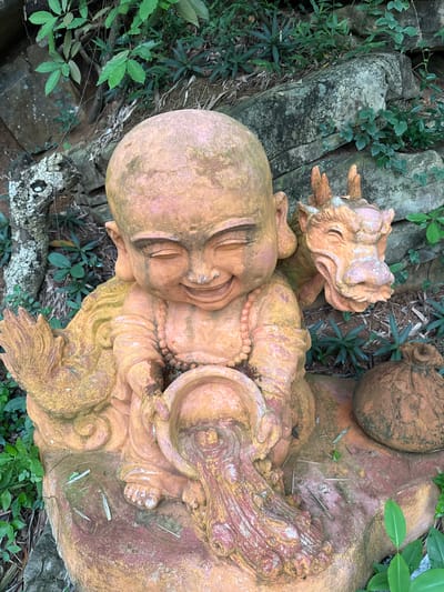 The image depicts a terracotta statue of a smiling child monk, seated with a small dragon perched on its right shoulder. The monk is holding a bowl from which a stream of water appears to be flowing. To the right of the monk, there is a sack-like object. The statue is situated outdoors, surrounded by rocks and lush green foliage, suggesting a garden or natural setting. The statue itself is weathered, with moss and dirt visible on its surface. The scene appears to be located in the city of Tây Hoa Lư Ward, Vietnam, a region known for its historical sites and natural beauty.