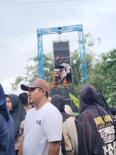 The image depicts an outdoor event or performance in Kerjo, Indonesia, under an overcast sky. In the center, a male performer stands atop a stack of large black audio speakers. He is dressed in a black long-sleeved shirt, a white, red, and black patterned loincloth or sarong, and red and yellow leg coverings. He holds a dark, shield-like object with red and black patterns. Above him, a speaker is suspended from a blue metal truss structure, which also supports the main speaker stack. Dense green foliage, including trees and bamboo, forms the background.
In the foreground, a crowd of spectators observes the event. A man in a white t-shirt and a brown patterned baseball cap is prominent on the left, looking towards the right of the frame. To his right, several individuals, primarily women, wear dark headscarves (hijabs). One person wears a black hooded sweatshirt with yellow and white text, including "NDX AKA," visible on the back. The crowd is positioned closely together, facing the performer and sound system.