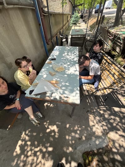 This is an eye-level, medium shot of four people gathered around a weathered outdoor table in Badiauri, Georgia. The scene is outdoors, shaded by trees, with dappled sunlight falling on the concrete ground and the table.

On the left, a woman with dark hair and glasses, wearing a yellow shirt, is eating. Next to her, another person's head and shoulders are visible, also with dark hair. To the right of the table, a teenage boy in a grey t-shirt and blue jeans is resting his head on his hand, looking down. Further to the right, another teenage boy in a black t-shirt is seated with his arms crossed, looking to the side.

The table is laden with bread, some of which is wrapped in paper. There are also plastic bags and a mobile phone on the table. The surroundings show a concrete wall, some pipes, a wooden bench, and in the background, a road with a white van and a black car. The overall impression is a casual gathering in a somewhat rustic outdoor setting.