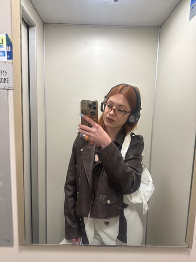 The image is a mirror selfie featuring a young woman with long, reddish-auburn hair, glasses with round dark frames, and black over-ear headphones. She is wearing a dark brown leather-style jacket over a white garment. Her visible hand, adorned with light blue nail polish, holds a smartphone with a patterned case, angled to capture her reflection.

The setting appears to be a small, enclosed interior space with plain white walls and ceiling, reflected in the mirror. Behind the woman's right shoulder (left in the reflection), a crumpled white object, possibly a bag or clothing, is visible.

The activity taking place is the woman posing for and taking a selfie in the mirror. Her expression is neutral, focused on her phone.

Notable details include the "MARSHAL" brand name visible on her headphones. On the bottom edge of the mirror's frame, reflected labels show partially legible text, including "315 SONE" and "FIAM," possibly referring to a manufacturer, which aligns with the Sanremo, Italy location context. The time of day and weather are not discernible.
