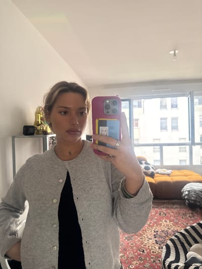 The image depicts a mirror selfie taken indoors within a residential setting in Nogent-sur-Marne, France. A female subject, appearing to be in her twenties or early thirties, is positioned centrally, holding a magenta-cased smartphone with her left hand, which features a clear cardholder containing a French student identification card. She has light-colored, wavy hair with highlights and is wearing a light grey, unbuttoned cardigan over a black top, revealing a small dark pendant on her neck. Multiple rings are visible on her left hand, and a dark green bracelet is on her left wrist.
The background, visible in the mirror's reflection, reveals a room furnished with various items. To the subject's left, a golden Buddha statue and a black JBL speaker rest on a light-colored shelf unit. To her right, a large window spans the wall, providing a view of multi-story residential buildings typical of an urban environment. Below the window, a mustard-yellow or orange-brown modular sofa or floor seating is present, adorned with a black and white cow print pillow and a white daisy-shaped cushion. The floor is covered by a patterned rug featuring red, orange, and dark motifs. The ceiling is plain white, with a single exposed light bulb fixture near the top center. The overall lighting suggests natural daylight entering through the window.