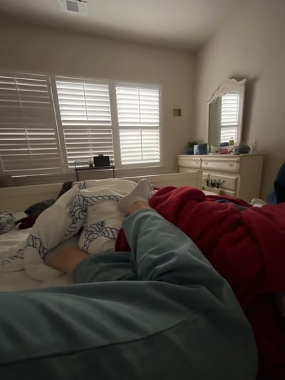 The image shows a cluttered bedroom interior. Two windows with white shutters are visible in the background, allowing natural light into the room. An air conditioning unit is installed on the wall near the left window. A white dresser is positioned against the wall, with various items on top, including what appears to be cleaning supplies. In the foreground, there are piles of bedding, including a red blanket, a white comforter with blue rope patterns, and possibly other items of clothing. A person is in the frame wearing a blue jacket.

There is no information in the image that indicates the location in the city of San Antonio, United States of America.