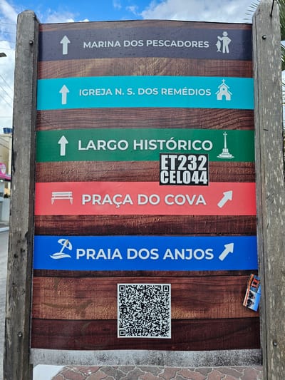 A vertical, rectangular directional sign, framed by two weathered, rough-hewn wooden posts, stands outdoors in Arraial Do Cabo, Brazil. The sign features five colored panels, each indicating a local landmark. From top to bottom:
1. A dark gray panel reads "MARINA DOS PESCADORES" with an upward arrow and an icon of a person holding a fish.
2. A teal panel reads "IGREJA N. S. DOS REMÉDIOS" with an upward arrow and a church icon.
3. A green panel reads "LARGO HISTÓRICO" with an upward arrow and a monument/cross icon. A black sticker with white text "ET232 CEL044" is affixed over the center of this panel.
4. A red panel reads "PRAÇA DO COVA" with a right-pointing arrow and a bench icon.
5. A blue panel reads "PRAIA DOS ANJOS" with a right-pointing arrow and an icon of a beach umbrella/palm tree.
Below the directional panels, a large black and white QR code is displayed on the brown, wood-grain patterned background of the sign. A small, folded brochure or sticker, displaying images of what appear to be boats or diving activities, is partially affixed to the right wooden post, slightly overlapping the lower right section of the sign's main face.
The sign is situated on a paved surface composed of reddish-brown interlocking pavers. In the background, under a partly cloudy blue sky, indistinct elements of an urban environment are visible, including power lines, a utility pole, and a building with a light-colored facade.