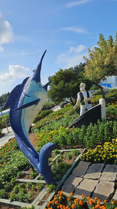 The image depicts an outdoor sculptural display in Cheokbeonjeong-ri, South Korea. In the foreground, a large, blue and white sculpture of a marlin is positioned diagonally, its body arched and tail fin raised. To the right and slightly behind the marlin is a sculpture of an elderly man with a white beard, wearing a black and white striped vest over a white shirt and pants. He is standing in a black boat and appears to be holding a rope. The sculptures are set within a garden landscape featuring abundant flowers, primarily in yellow and orange hues, interspersed with lush green foliage. Stone pathways are visible in the lower right portion of the image. In the background, trees are present, along with structures that appear to be part of a greenhouse or modern building, visible on the far right. The sky above is blue with scattered white clouds.
