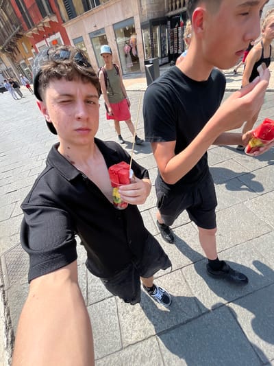 The image presents a first-person perspective snapshot captured on the streets of Verona, Italy. Two young individuals are holding cups of mixed fruit, possibly watermelon and other fruits, on skewers. Both are dressed in dark clothing and shoes, with one wearing shorts and the other dressed in a black button-down shirt. The background reveals the architectural landscape typical of Verona, including shops with displays of goods and other pedestrians strolling by. The shot is taken on a sunny day, casting shadows on the paved street.