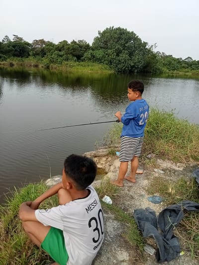 Two young boys are fishing by a calm body of water, likely a river or lake, in Teluk Kenidai, Indonesia. The boy on the right, wearing a blue t-shirt with "PALMER 20" on the back and striped shorts, stands with a fishing rod in his hands. The boy on the left, wearing a white t-shirt with "MESSI 30" on the back and green shorts, is sitting on the ground facing away from the camera, also appearing to be fishing. The environment is lush and green, with dense vegetation along the banks of the water. The sky is overcast, suggesting a cloudy day. The ground is uneven, with patches of grass and exposed soil.