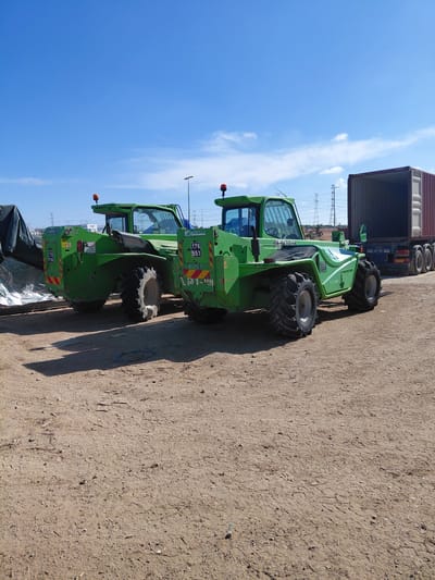 Two green Merlo telehandlers are parked on a dirt lot in Caesarea, Israel. A shipping container is visible in the background, along with power lines. The sky is blue with some clouds.