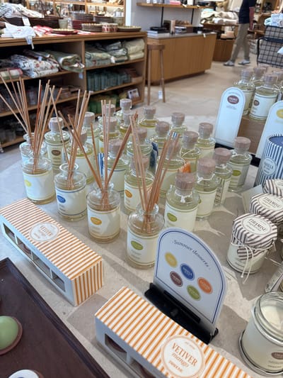 The image shows a display of home fragrance products, likely in a retail store. In the foreground, there are several glass bottles with wooden stoppers and reeds, indicating reed diffusers. These bottles have labels with various scent names, such as "VETIVER MANGO," "VANILLA & NUT," and "TIRAMISU EUPHORIA." A box with an orange and white striped pattern is also visible, labeled "VETIVER MANGO scented pillar candle." To the right, there are jars with striped lids, also labeled with scent names. A sign with the heading "Summer desserts" displays several oval labels corresponding to different scents. In the background, shelves are stocked with various home goods, including linens and tableware. The lighting suggests an indoor retail environment. The city of Orihuela, Spain, is not depicted or implied by any elements in the image. There are no people clearly visible in a way that allows for detailed description. The overall scene is one of organized product display within a store.