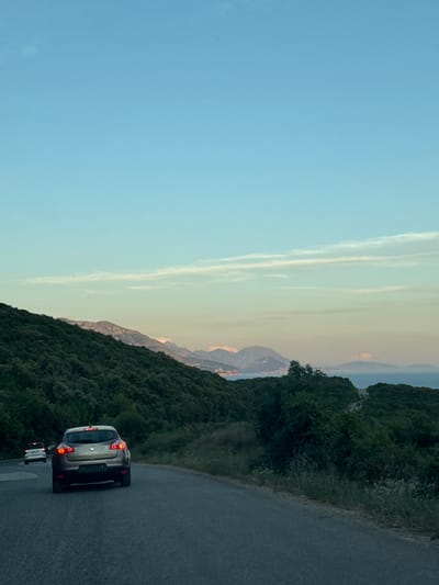 An asphalt road winds through a lush, green landscape under a clear blue sky with wispy clouds. The road curves to the left, with dense foliage and trees lining its right side. In the foreground, a dark-colored sedan, possibly a Renault, is driving away from the camera. Its brake lights are illuminated, indicating deceleration. Further ahead on the road, a white car is also visible, moving in the same direction. In the background, distant mountains are silhouetted against the horizon, with a body of water, likely the sea, and scattered islands visible between the mountains and the coast. The overall impression is of a scenic drive along a coastal route, possibly in the vicinity of Kubasi, Montenegro, during the late afternoon or early evening.
