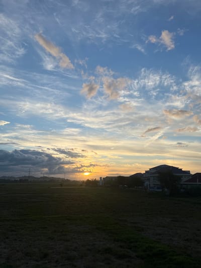 The image depicts a landscape scene in the city of Meureudu, Indonesia, during what appears to be sunset or sunrise. A vast expanse of dark, mowed grass dominates the foreground, indicating a field or park. In the middle ground, a line of trees and buildings runs horizontally across the frame. Among these structures are several multi-story buildings, suggesting a residential or commercial area. Power lines and transmission towers are visible in the distance, hinting at the presence of infrastructure. The sky is a prominent feature, filled with a variety of clouds. Wispy cirrus clouds streak across a bright blue sky in the upper portion, while lower, more textured clouds are tinged with the warm hues of the setting or rising sun. The sun itself is visible as a bright, golden orb peeking through the clouds on the right side of the frame, casting a warm glow across the sky and horizon. The overall impression is one of a serene, natural environment contrasted with man-made structures. There are no people or animals visible in the image.