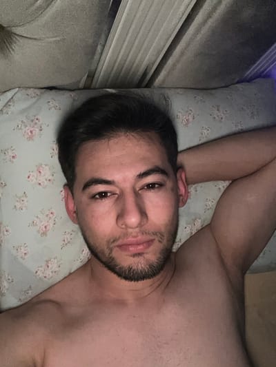 A bare-chested man is lying on a pillow with a floral print. He is looking directly at the camera, his head tilted slightly. He has dark hair and a short beard. His arm is raised above his head. A window with white blinds is visible to the left of the frame. The overall lighting is dim.